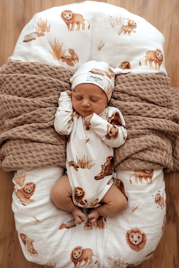 Newborn baby in long-sleeve bodysuit printed with bronze lion designs on white, with a matching beanie laying on a caramel knitted blanket and lion snuggle bed at Little Ellie Boutique, an Australian baby shop