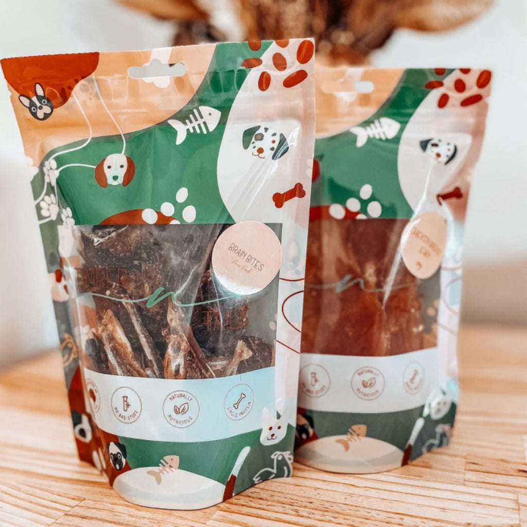 Two bags of organic, dehydrated dog treats in green and white eco-friendly packaging on a wood table.