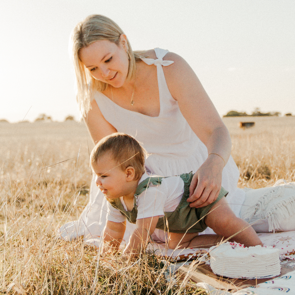 Seamstress for That's Sew Ange, in a white dress kneels over baby in white shirt and olive green frilled romper in grassy paddock, available at Little Ellie Boutique, an Australian baby shop