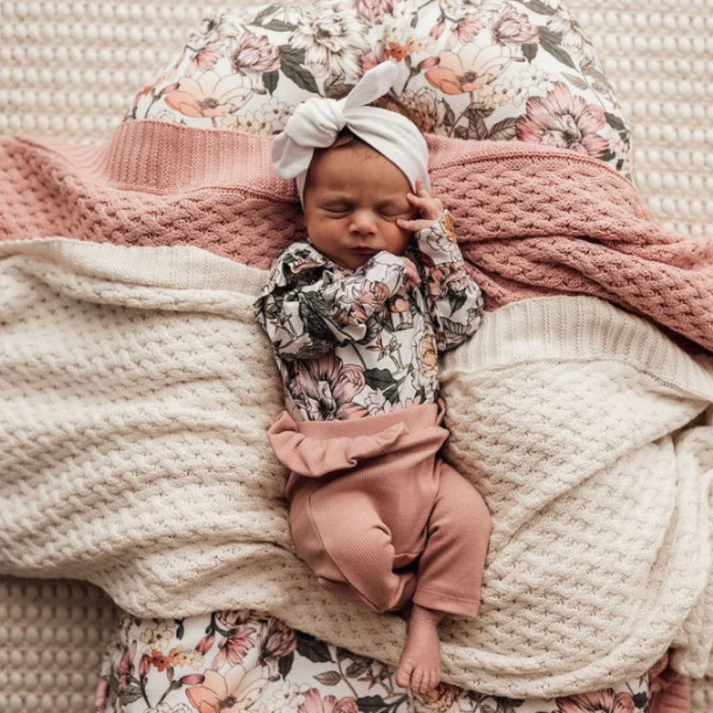 A newborn baby dressed in frilled pink ribbed pants and floral jersey bodysuit topped with a knotted headband lays on a knitted cream and pink blanket, from Little Ellie Boutique, an Australian baby shop