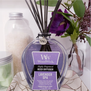Purple lavender spa woodwick reed diffuser sits on a gold and pink marble plate with home decor spaced behind it.